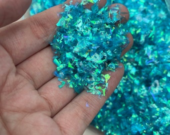 Könnte beinhalten: Nahaufnahme einer Hand, die einen Haufen schimmernder türkisfarbener Glitzerflocken hält. Der Glitzer befindet sich in einem durchsichtigen Plastikbeutel.