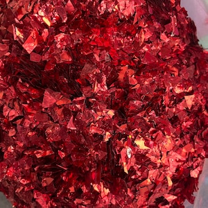 May include: A close-up of a large pile of red glitter confetti. The confetti is made of small, irregular pieces of shiny red foil.