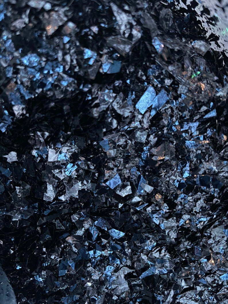 May include: A close-up of a pile of dark blue iridescent glass pieces. The glass is broken into small, irregular shapes and has a shimmering, reflective surface.