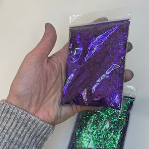Op de afbeelding: Twee kleine zakjes met iriserende glitter. Eén zakje bevat paarse glitter en het andere groene glitter.