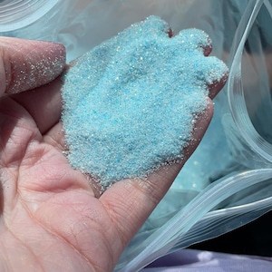 Może przedstawiać: Zbliżenie na dłoń trzymającą woreczek z jasnoniebieskim brokatem. Brokat znajduje się w przezroczystym plastikowym woreczku z zamkiem błyskawicznym.