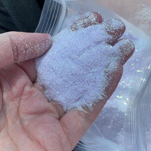 Puede incluir: Primer plano de una mano sosteniendo una bolsa de purpurina blanca iridiscente. La purpurina es muy fina y tiene un ligero tono púrpura.
