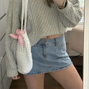 May include: A white crochet bag with a pink bow is held in front of a person wearing a light grey cable knit sweater and a denim mini skirt. The bag has a long strap and a cylindrical shape.