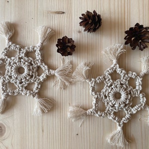 May include: Two beige crocheted snowflakes with tassels on a wooden surface. The snowflakes are made with a macrame technique and have a delicate, intricate design.