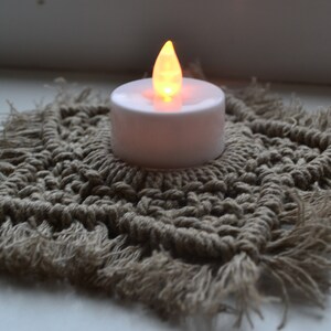 May include: A small, white, battery-operated candle with a warm, orange glow sits on a beige crocheted coaster with fringe.