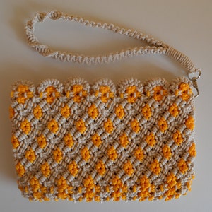 May include: A beige macrame clutch with a yellow zigzag pattern and a matching wrist strap. The clutch has a scalloped edge and a silver-tone clasp. The handmade bag is a stylish accessory.