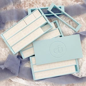 May include: A collection of light blue gift boxes with white inserts, each bearing embossed text for various occasions like "Bridal Shower" and "Happy Anniversary." The boxes are arranged on a white lace surface with blue ribbons.