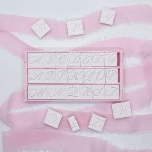 May include: A pink plastic stamp set with white letter stamps in a cursive font. The set includes a tray with the alphabet, plus individual letter stamps and a pink ribbon on a white background.