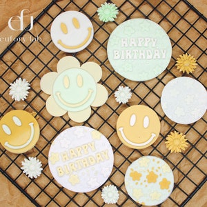 Könnte beinhalten: Eine Sammlung dekorativer Smiley-Gesichter und blumenförmiger Designs in Pastellgelb, Grün und Weiß. Einige Designs sind rund mit einem Smiley-Gesicht, andere blumenförmig oder mit der Aufschrift "HAPPY BIRTHDAY". Die Artikel sind auf einem schwarzen Drahtgestell angeordnet.