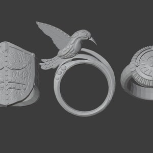 Puede incluir: Tres anillos de plata con diferentes diseños. Un anillo presenta un escudo con detalles intrincados, otro tiene un diseño de colibrí y el tercero tiene un diseño circular con un patrón de rayos de sol.