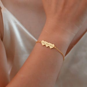 Op de afbeelding: Een goudkleurige armband met drie hartvormige bedels. Elke bedel heeft een gegraveerde naam. De armband wordt om een pols gedragen. De achtergrond is een zachte, neutrale kleur.
