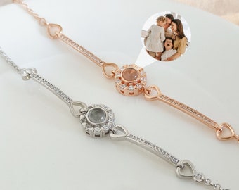 Puede incluir: Dos pulseras delicadas, una plateada y otra de oro rosa, cada una con un dije circular central para la proyección de fotos. Las pulseras tienen detalles en forma de corazón y están adornadas con pequeños cristales. El fondo es una superficie blanca y suave.