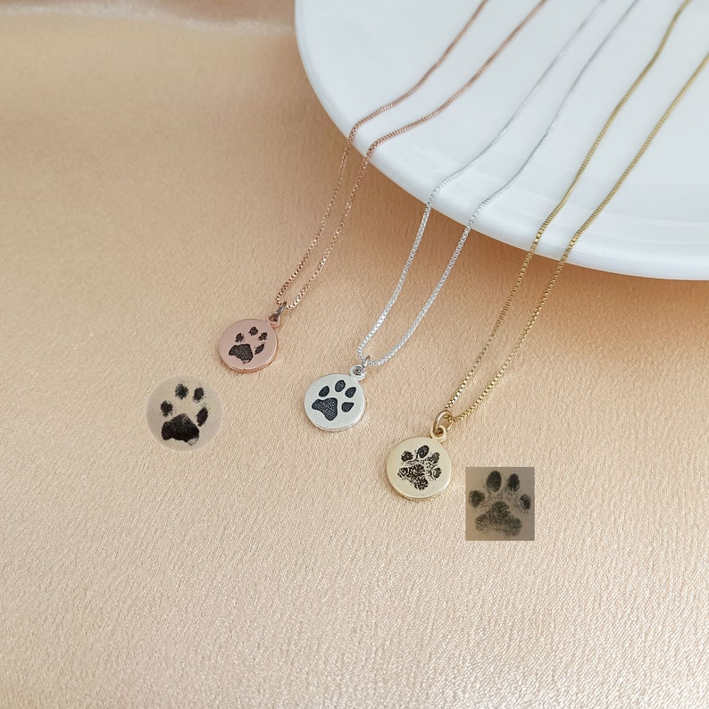 May include: Four necklaces with silver, gold, and rose gold chains and paw print pendants. The pendants are engraved with a paw print.