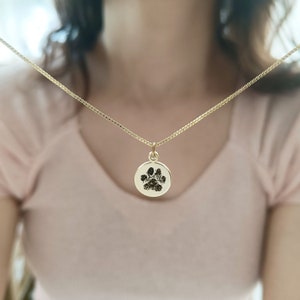 May include: A gold necklace with a round pendant featuring a black paw print. The necklace is worn by a person with long dark hair.