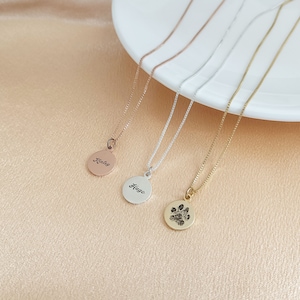 May include: Four gold necklaces with different pendants. The pendants are a round disc with the name "Riley" engraved on it, a round disc with the name "Hugo" engraved on it, and a round disc with a paw print engraved on it.