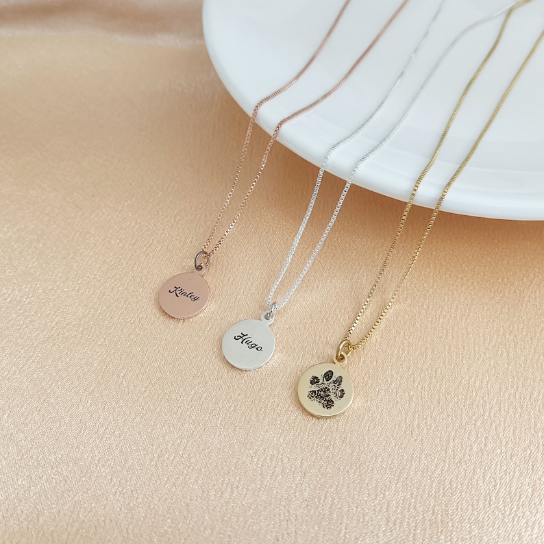 May include: Four gold necklaces with different pendants. The pendants are a round disc with the name "Riley" engraved on it, a round disc with the name "Hugo" engraved on it, and a round disc with a paw print engraved on it.