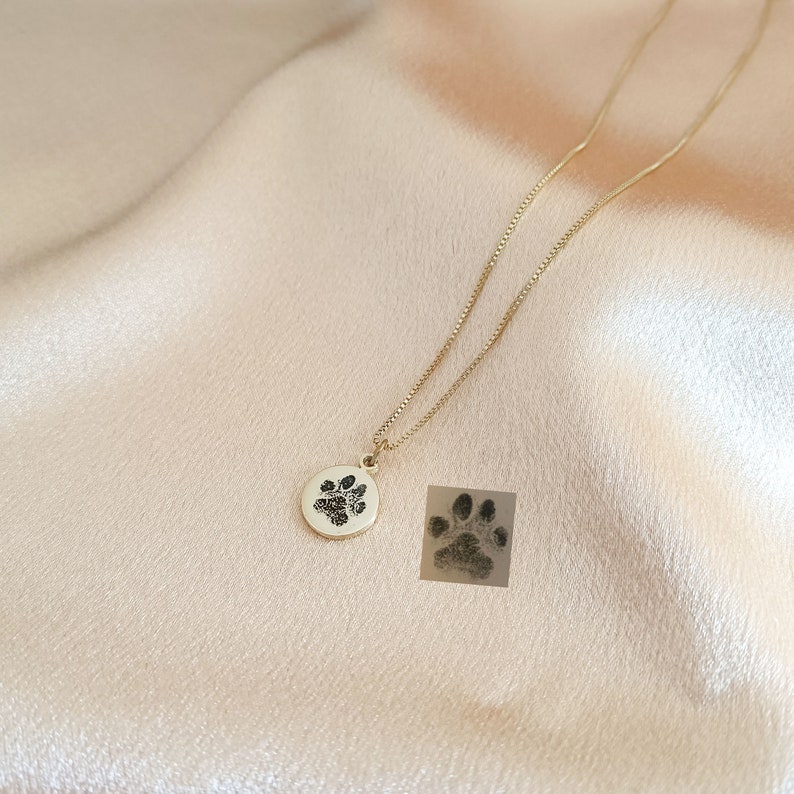 May include: A gold necklace with a round pendant featuring a black paw print. The pendant is on a delicate chain.