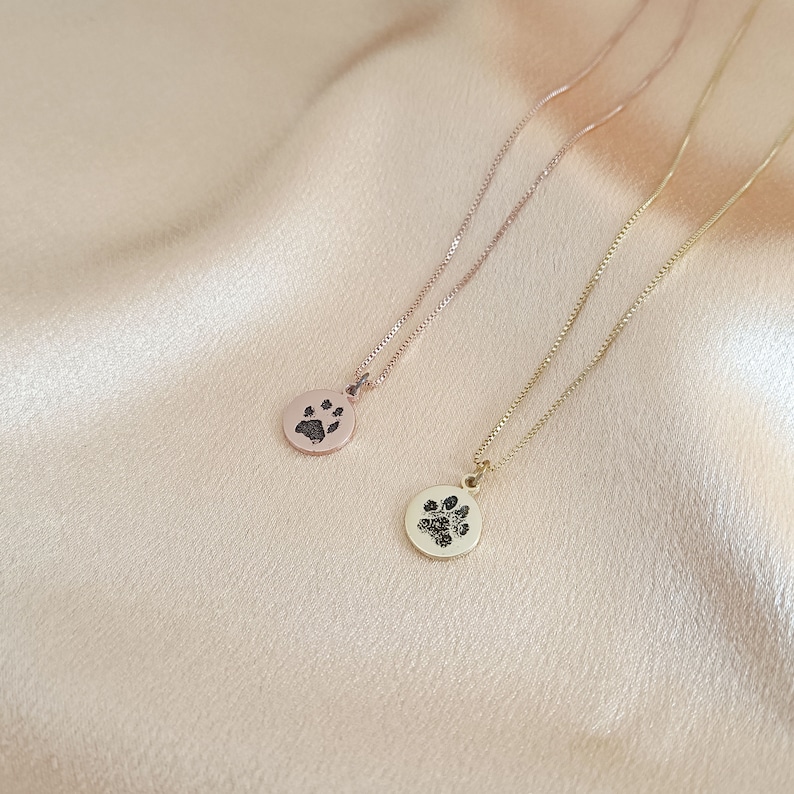 May include: Two gold necklaces with paw print pendants. One necklace has a rose gold chain and the other has a yellow gold chain.