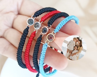 Puede incluir: Tres pulseras de la amistad con un dije de foto. Las pulseras son rojas, negras y azules. El dije de foto es un círculo de vidrio transparente con una foto de una familia dentro.