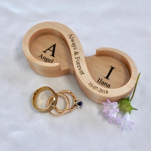 Puede incluir: Soporte de anillos de madera con forma de infinito, grabado con "Always & Forever", nombres y una fecha. Incluye tres anillos dorados y un pequeño ramo de flores moradas. El soporte es de madera clara.
