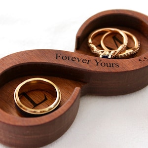 May include: A wooden ring box shaped like an infinity symbol. The box is engraved with the words "Forever Yours" and the date "5-5-2018". Two gold rings are inside the box.