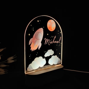 May include: A decorative night light featuring a rocket ship, moon, and clouds against a starry black background. The name "Michael" is written in a cursive font. The light has a wooden base and an arched top.