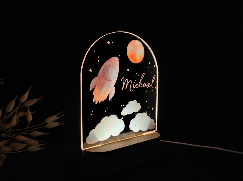 May include: A decorative night light featuring a rocket ship, moon, and clouds against a starry black background. The name "Michael" is written in a cursive font. The light has a wooden base and an arched top.