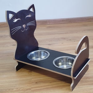 Könnte beinhalten: Ein schwarzer Futterständer aus Holz mit einem Katzen-Silhouetten-Design. Der Ständer hat zwei Edelstahlschalen für Futter und Wasser.