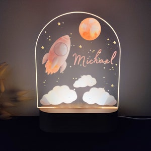 May include: A decorative night light featuring a rocket, moon, and clouds design. The light is personalized with the name "Michael" in pink script. The lamp has a wooden base and a clear acrylic front.