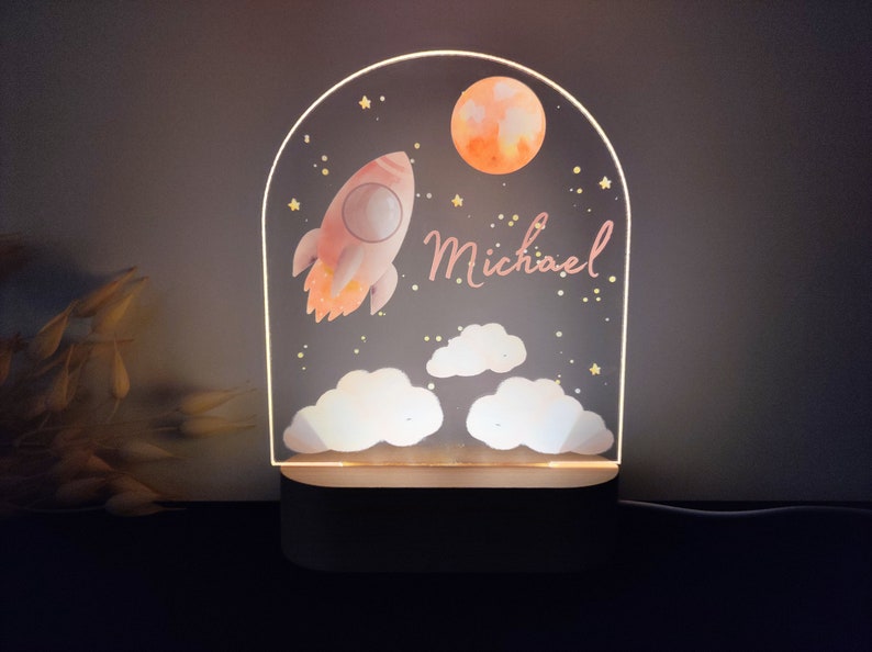 May include: A decorative night light featuring a rocket, moon, and clouds design. The light is personalized with the name "Michael" in pink script. The lamp has a wooden base and a clear acrylic front.