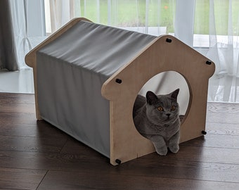 Può includere: Un gatto grigio con occhi gialli è seduto dentro una cuccia per gatti in legno con un tetto in tessuto grigio. La cuccia ha un'apertura rotonda nella parte anteriore.