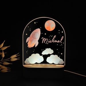 May include: A decorative night light featuring a rocket ship, moon, stars, and clouds against a black background. The name "Michael" is written in cursive. The light has a wooden base and a glowing acrylic panel.