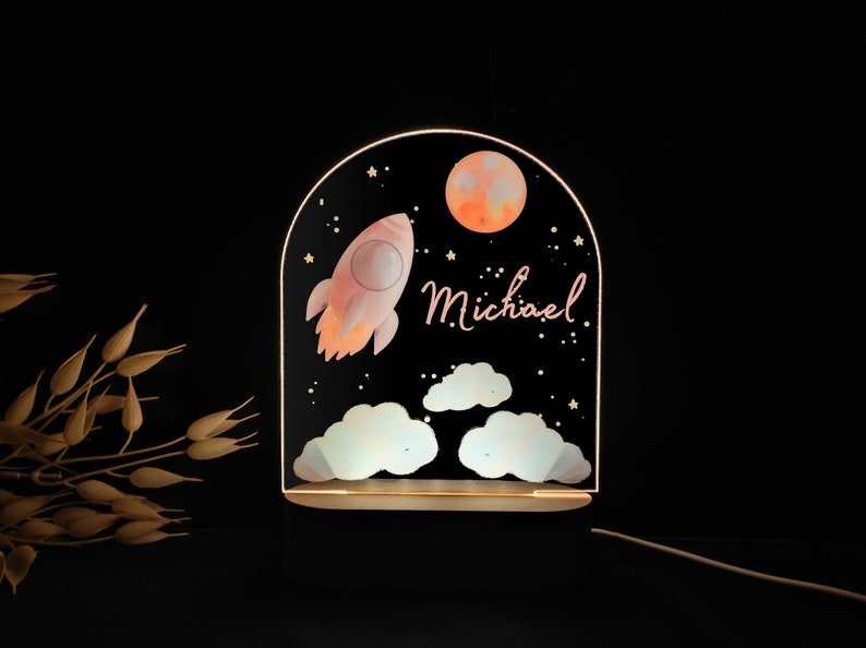 May include: A decorative night light featuring a rocket ship, moon, stars, and clouds against a black background. The name "Michael" is written in cursive. The light has a wooden base and a glowing acrylic panel.
