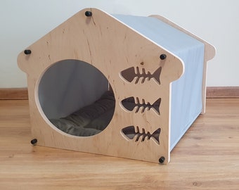 Può includere: Una cuccia per gatti in legno con un tetto in tessuto grigio e un'apertura rotonda. La cuccia ha tre tagli a forma di lisca di pesce sul davanti.