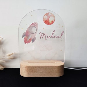 May include: A personalized night light featuring a pink rocket, a red moon, and the name "Michael" in script. The design is printed on a clear acrylic panel, set on a wooden base. The light is powered by a white cord.