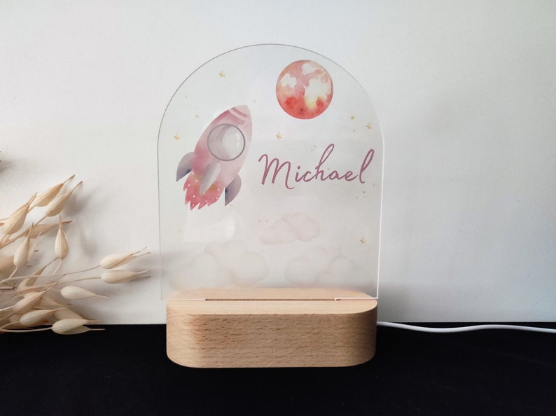 May include: A personalized night light featuring a pink rocket, a red moon, and the name "Michael" in script. The design is printed on a clear acrylic panel, set on a wooden base. The light is powered by a white cord.