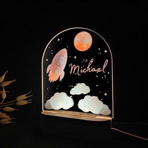 May include: A decorative night light featuring a rocket, moon, clouds, and stars against a black background. The name "Michael" is written in cursive. The light is set on a wooden base and emits a warm glow.