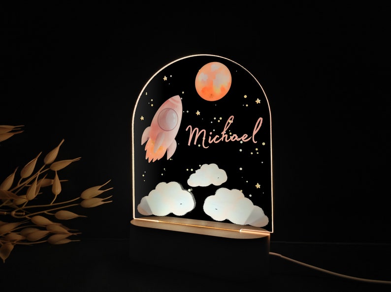 May include: A decorative night light featuring a rocket, moon, clouds, and stars against a black background. The name "Michael" is written in cursive. The light is set on a wooden base and emits a warm glow.
