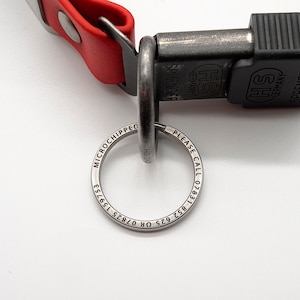 May include: Close-up of a red leather dog collar with a black clasp and a silver key ring. The key ring has the words "MICROCHIPPED" and "PLEASE CALL" with a phone number. The clasp is marked "SPRENGER GERMANY".