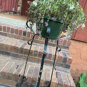 May include: A dark green hanging planter filled with trailing plants and flowers, supported by a black wrought iron plant stand. The stand has decorative scrollwork and is placed on brick steps.