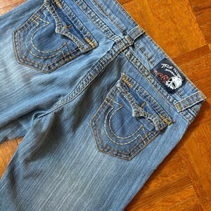 May include: Light blue denim jeans with a faded wash. The back pockets feature a distinctive horseshoe design stitched in gold thread. The True Religion brand label is visible at the waistband. The jeans have gold stitching.