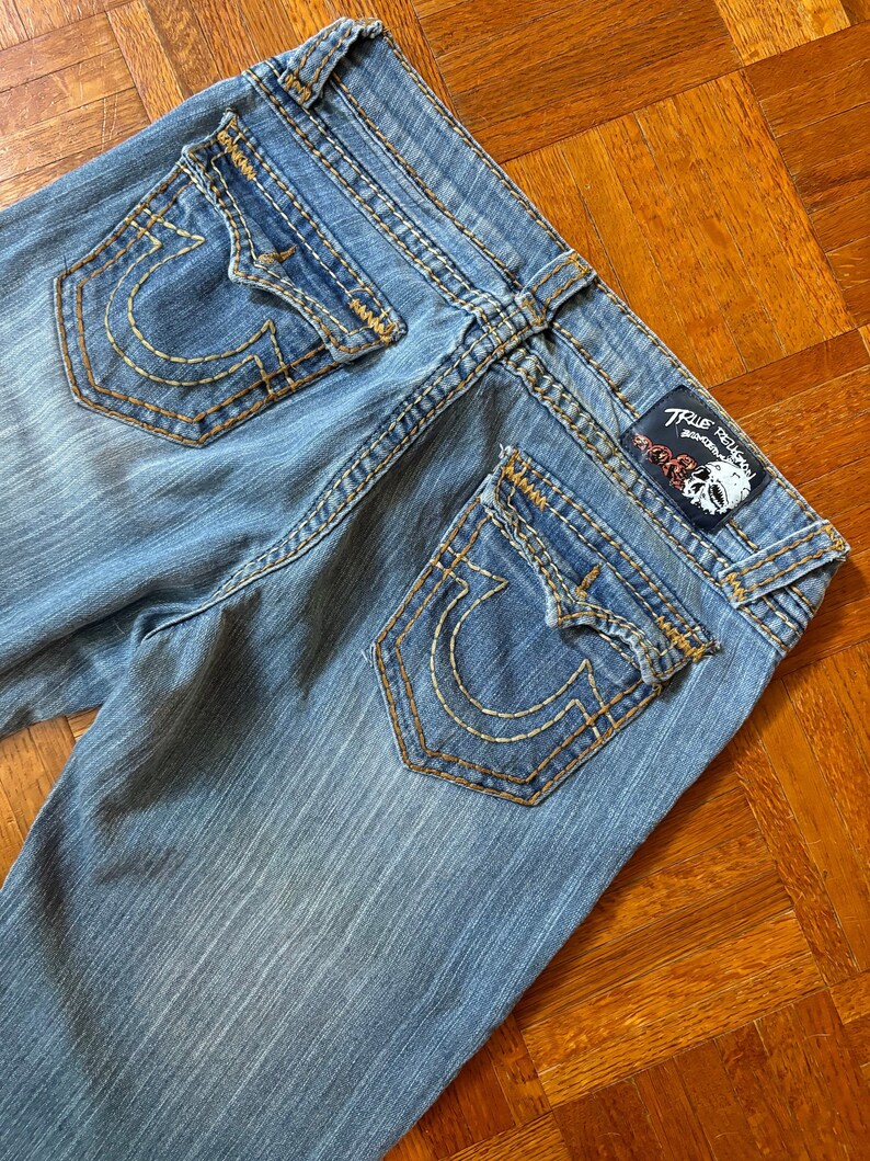 May include: Light blue denim jeans with a faded wash. The back pockets feature a distinctive horseshoe design stitched in gold thread. The True Religion brand label is visible at the waistband. The jeans have gold stitching.