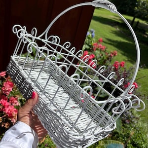 May include: White wicker basket with a decorative handle and scrollwork design. The basket has a rectangular shape and is painted white. The handle is arched and attached to the basket with a twisted design. The basket is held in front of a background of flowers and greenery.
