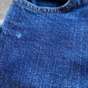 Op de afbeelding: Close-up van blauwe denim stof met een klein gaatje. De stof heeft een textuur en een subtiele glans.