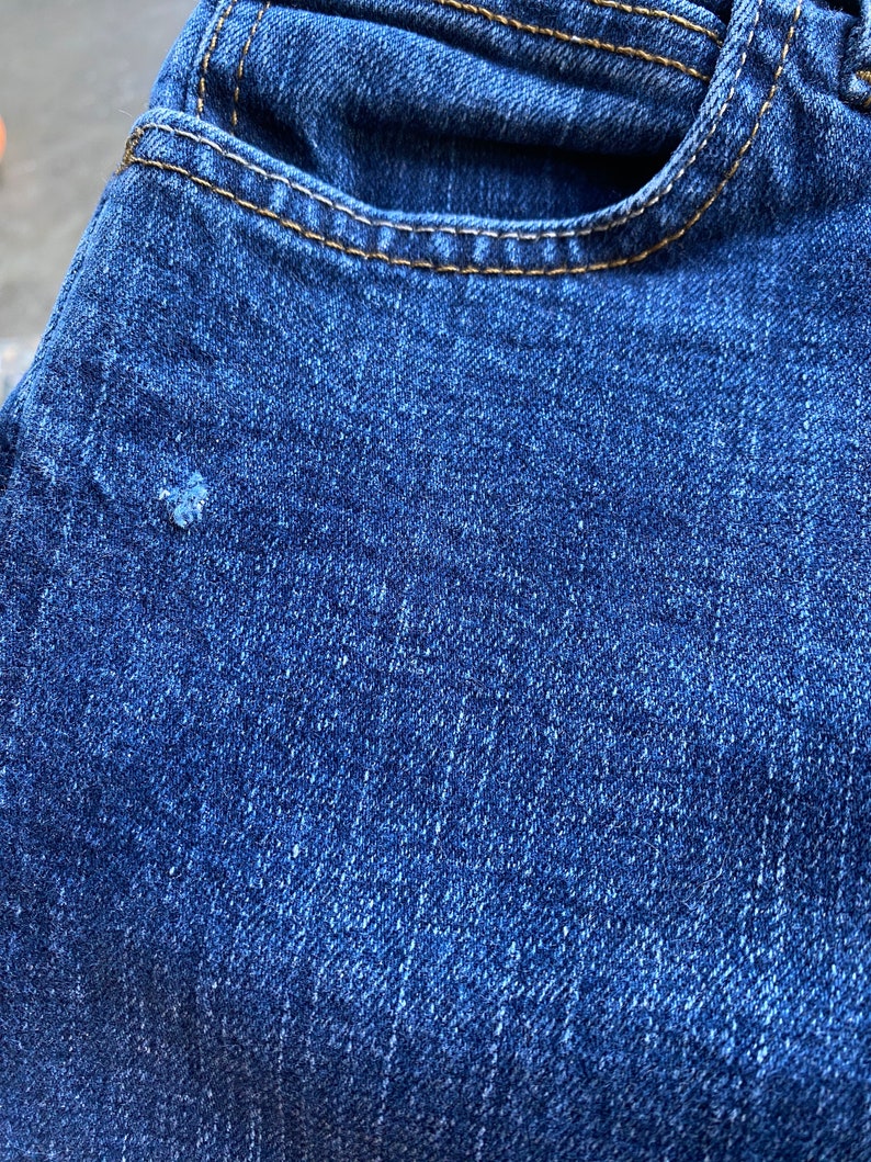 Op de afbeelding: Close-up van blauwe denim stof met een klein gaatje. De stof heeft een textuur en een subtiele glans.