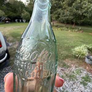 Puede incluir: Una botella de Coca-Cola vintage de vidrio verde claro con forma contorneada. El texto en relieve en el frente dice Coca-Cola, Registered y 6 FL OZS. La botella presenta un cuello estrecho y una base ancha con estr&iacute;as verticales.