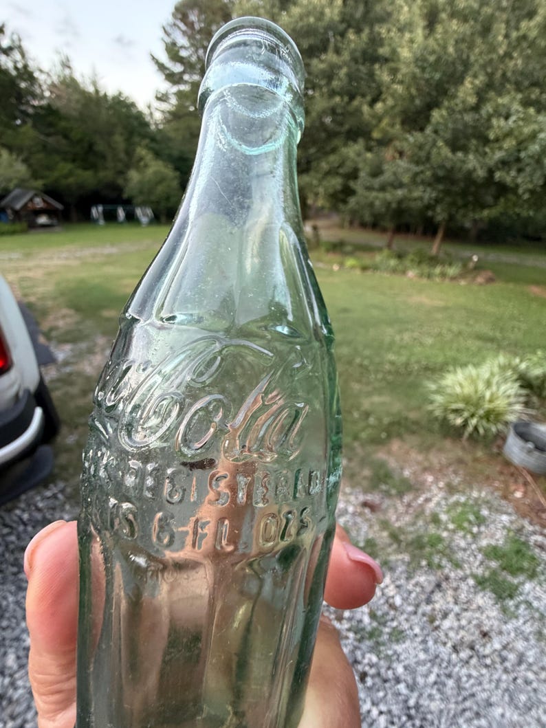 Puede incluir: Una botella de Coca-Cola vintage de vidrio verde claro con forma contorneada. El texto en relieve en el frente dice Coca-Cola, Registered y 6 FL OZS. La botella presenta un cuello estrecho y una base ancha con estr&iacute;as verticales.