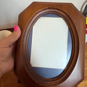 May include: A medium-toned wooden picture frame with an octagonal outer shape and a central oval opening. The frame features a smooth, polished finish and contains a dark blue mat board surrounding a rectangular white insert for a photograph.