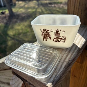 Puede incluir: Plato cuadrado de vidrio blanco con un diseño marrón de maíz, una estufa y un gato. El plato tiene una tapa de vidrio transparente con nervaduras.