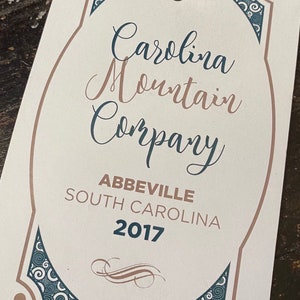 May include: A white tag with brown and blue decorative borders. The tag has the text "Carolina Mountain Company" in cursive script, followed by "Abbeville, South Carolina 2017".